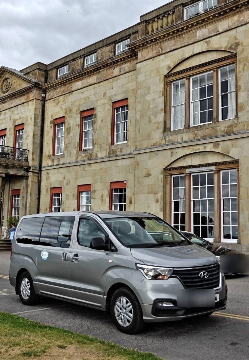 A silver minibus on the drive of a large country house.