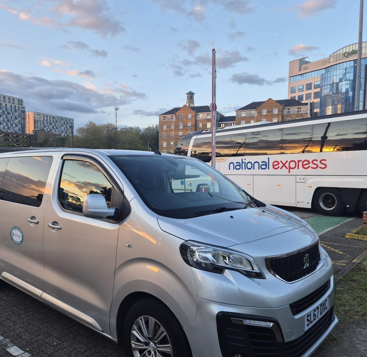 A silver minibus parked at a coach station at golden hour, a coach behind it.