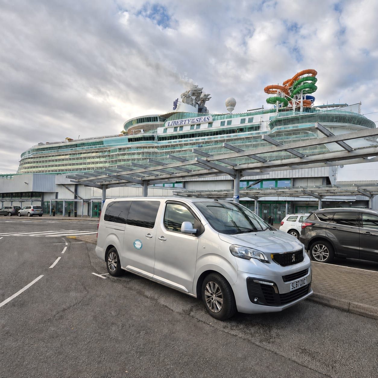 A silver minibus at a cruise terminal with a cruise ship berthed behind it.
