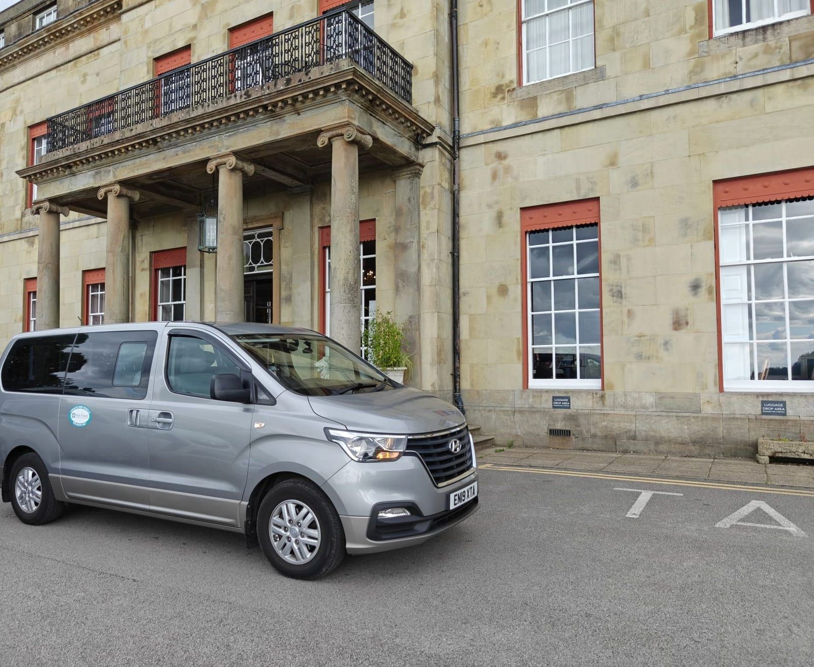 A silver minibus outside the stone frontage of a large country house.
